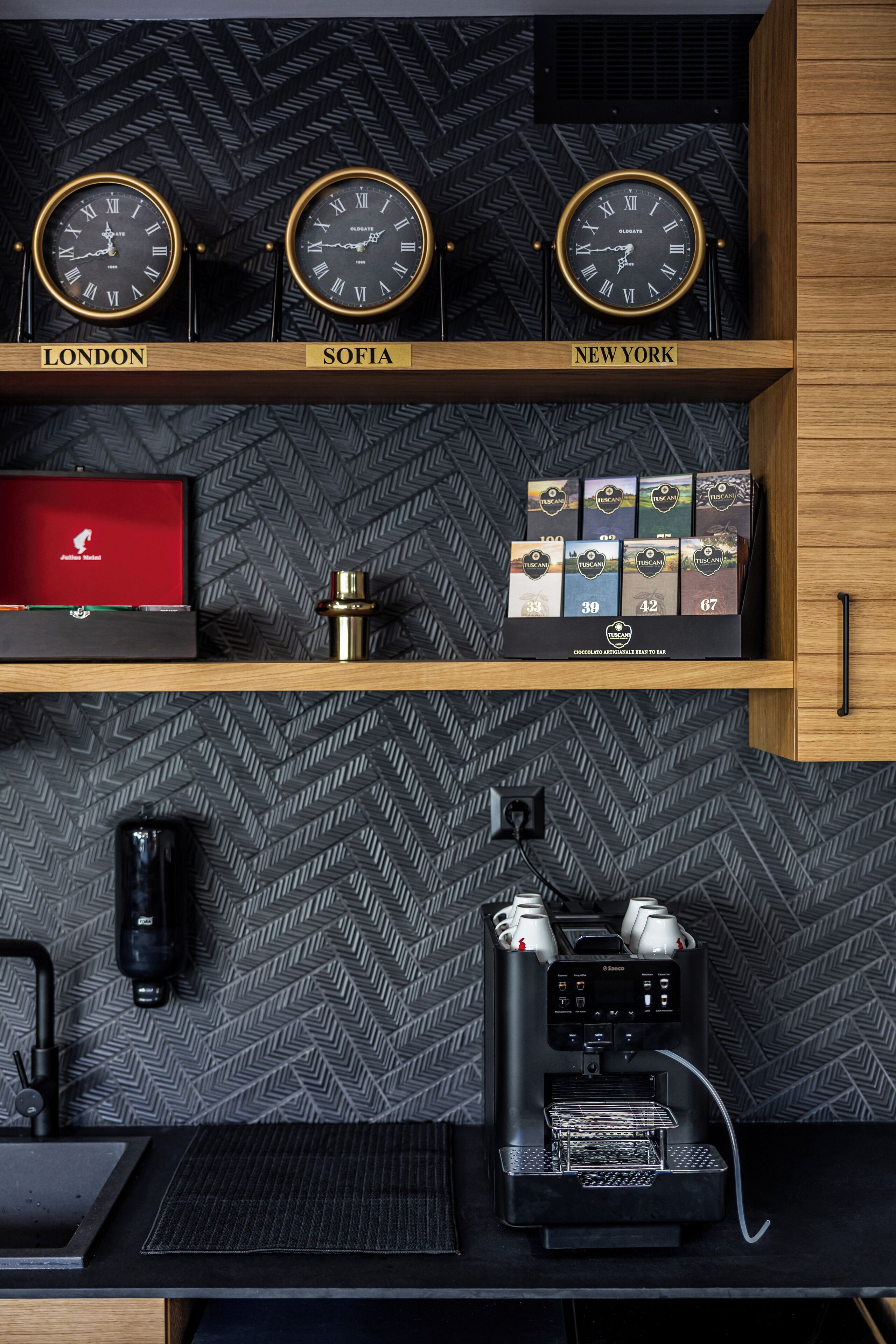 Een muur met drie klokken met de aanduiding London, Sofia en New York, een plank met dozen vol Toscaanse koffie, en een koffieapparaat.