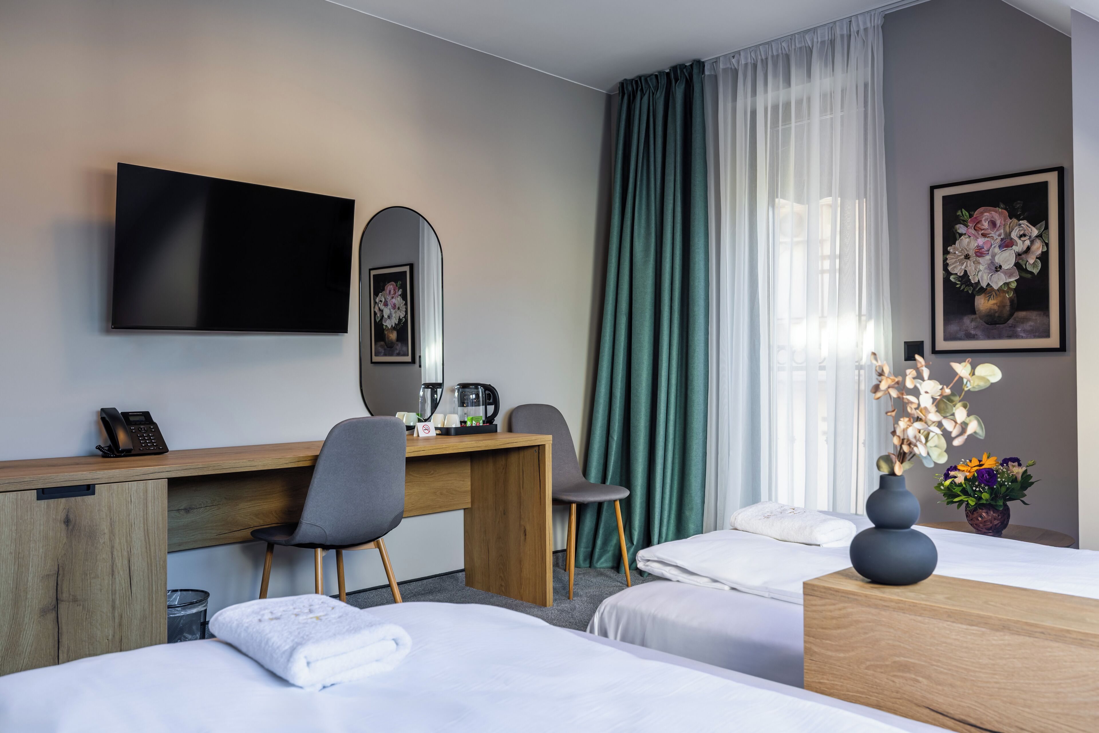 A hotel room with two beds, a desk with a chair, a TV, a coffee maker, and a vase with flowers.