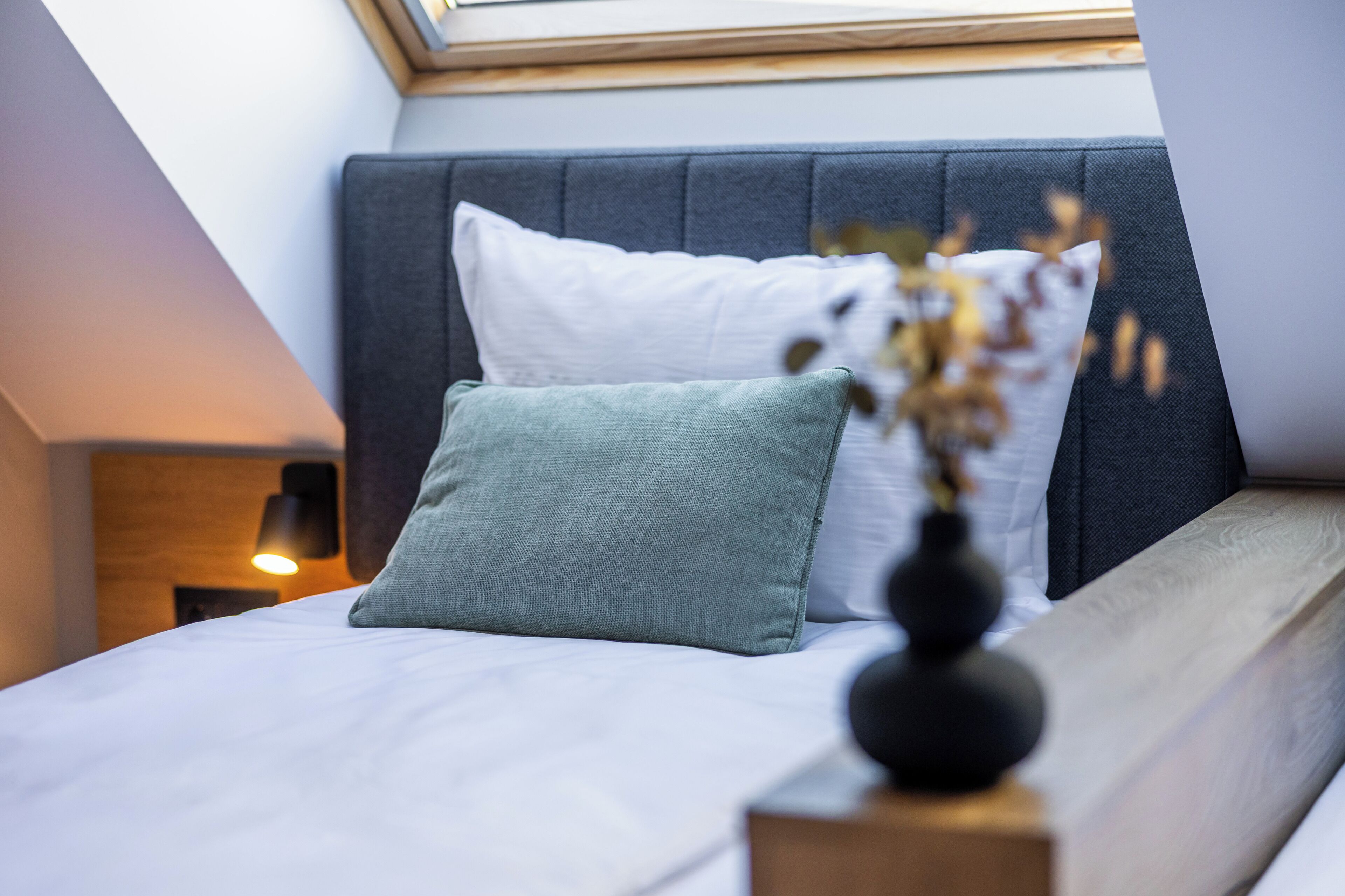A neatly made bed with two pillows, a bedside table with a vase, and a nightstand with a lamp.