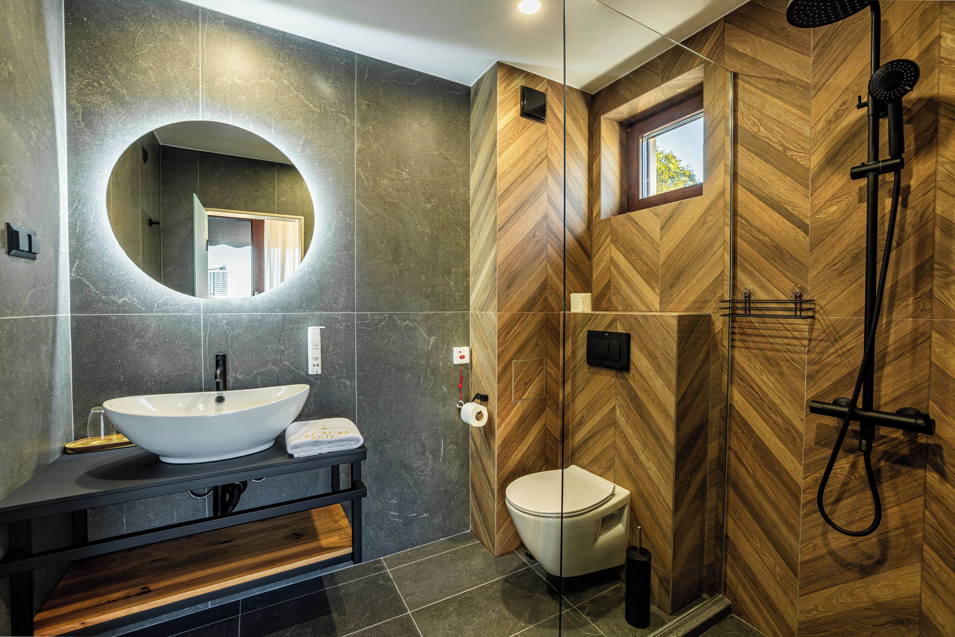 A modern bathroom with a large oval mirror, a vessel sink, a toilet, and a walk-in shower with wooden paneling.