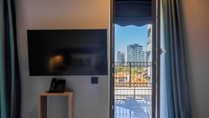 A room with a mounted TV, a telephone, and a view of a cityscape through a window with a metal railing. - Acacia Boutique Hotel (Sofia)