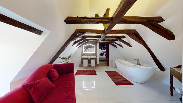 An attic bedroom with a sloped ceiling, a large window, a red sofa, a white bathtub, and a mirror above the sink. - Château Ecrin de lumiere (Campsegret)