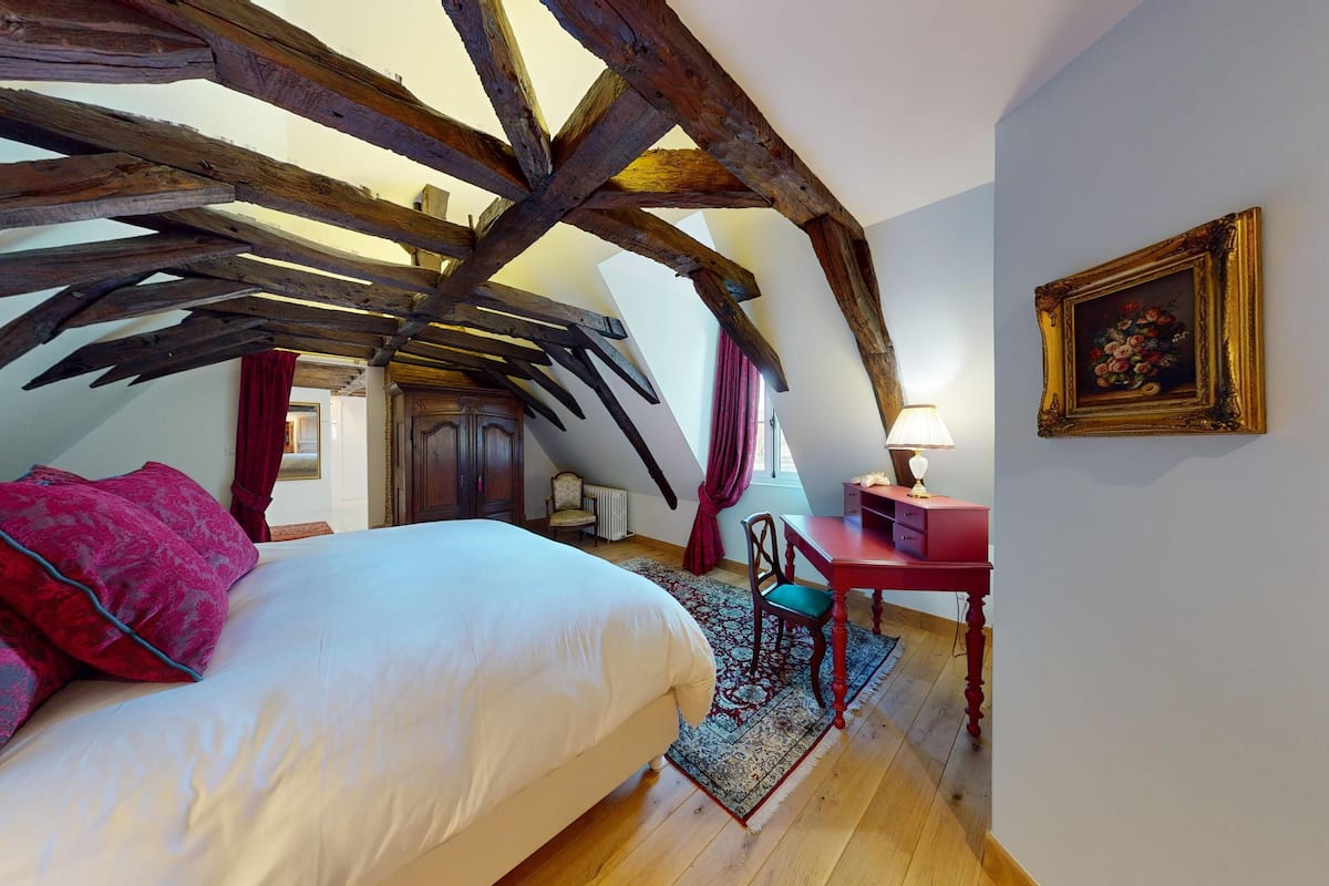 A bedroom with a sloped ceiling, a bed with red and white bedding, a wooden desk with a lamp, and a framed painting on the wall.