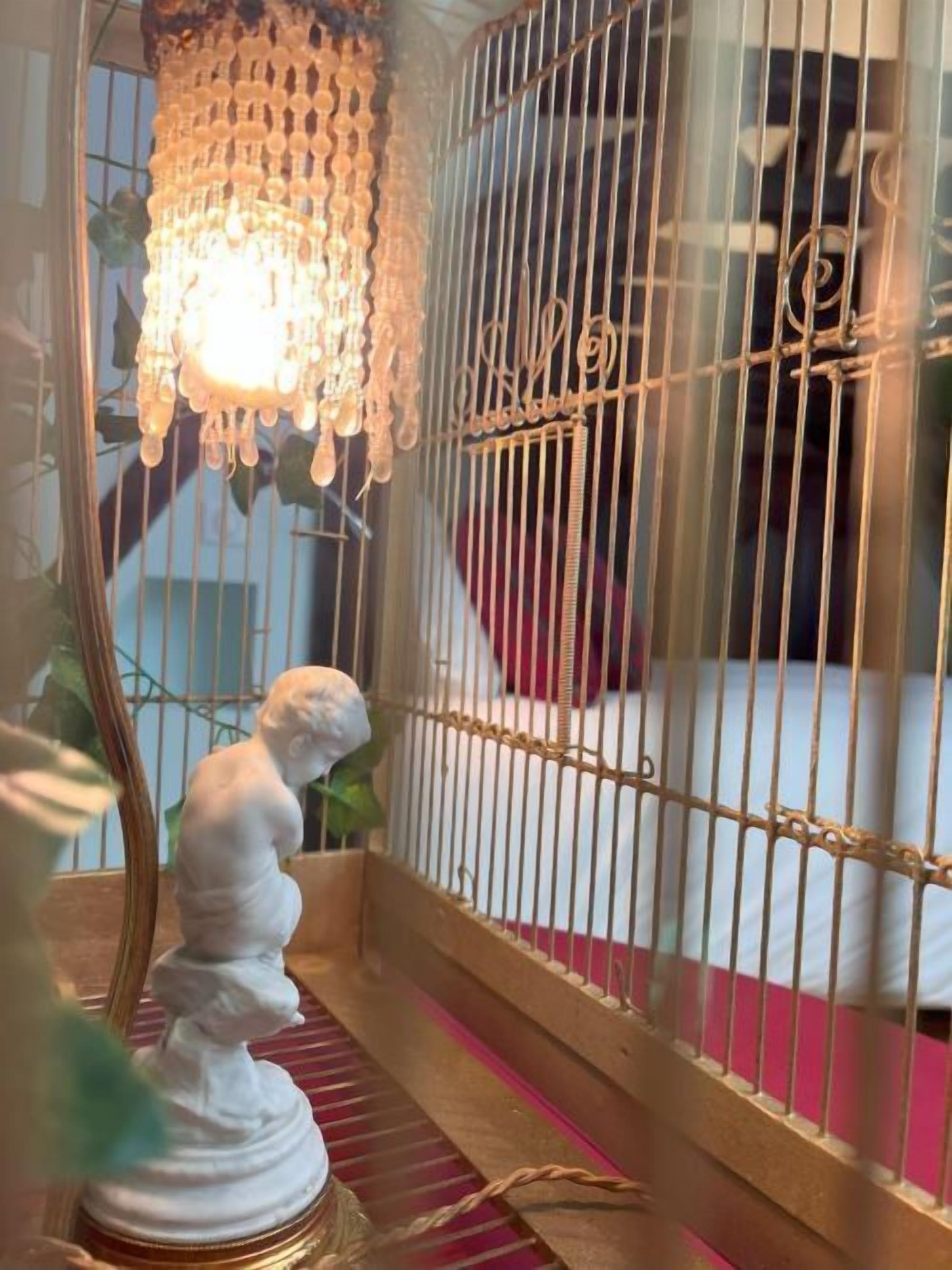 A birdcage with a decorative hanging light fixture, a white statue, and a bed with a red pillow in the background.