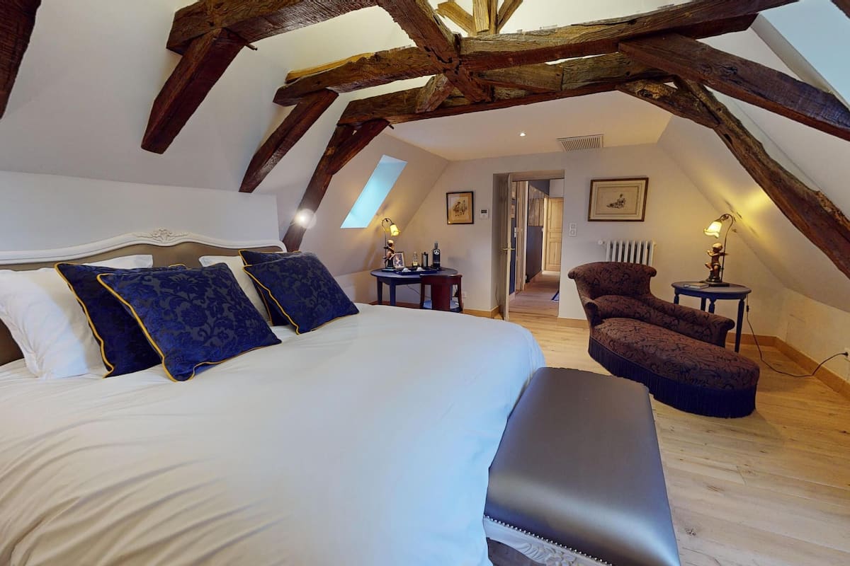 A bedroom with a large bed, a chair, and a desk. There is a skylight and wooden beams on the ceiling.