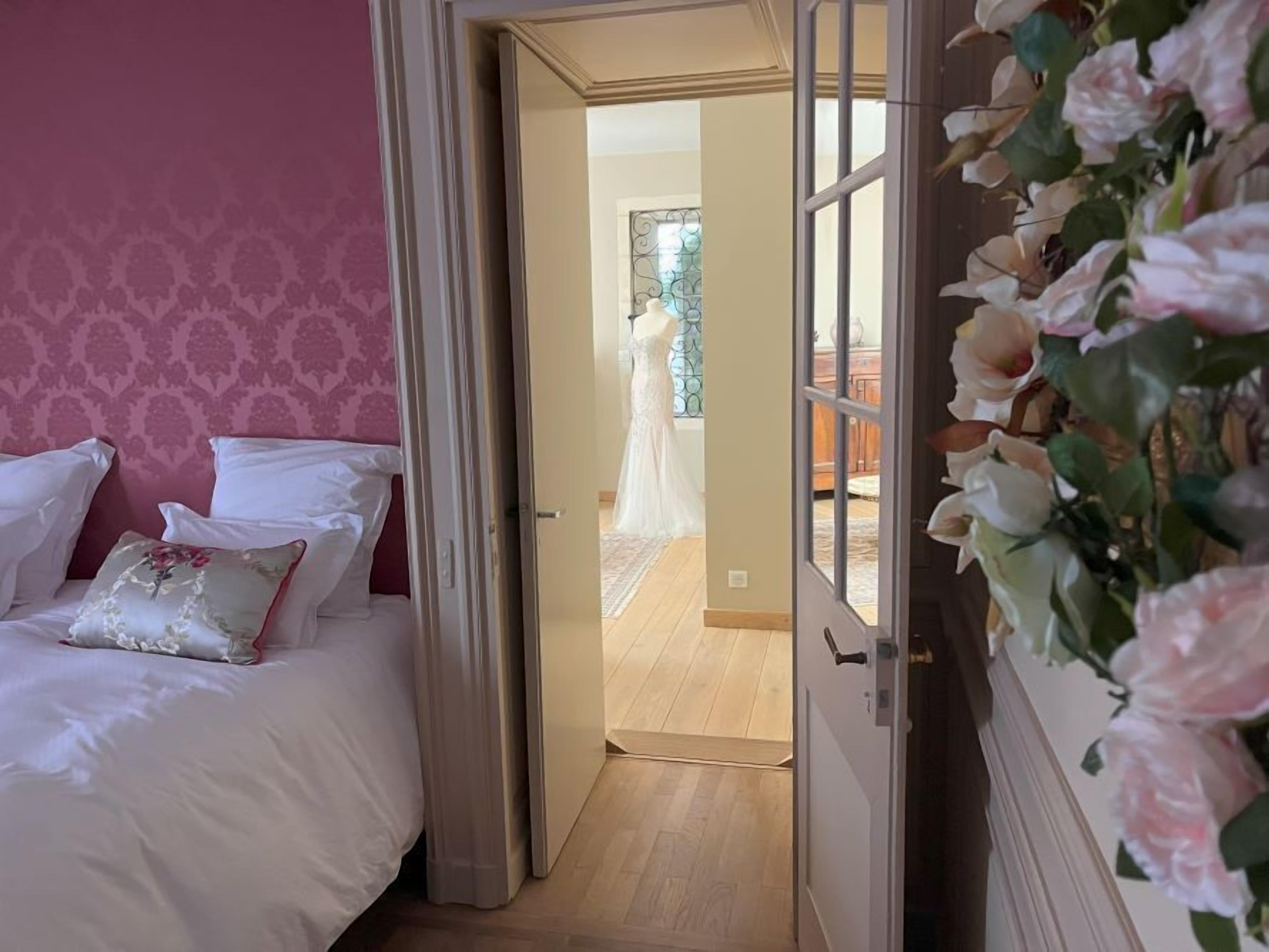 A bedroom with a bed, pillows, a door leading to a room with a person in a white dress, and a window with a view.