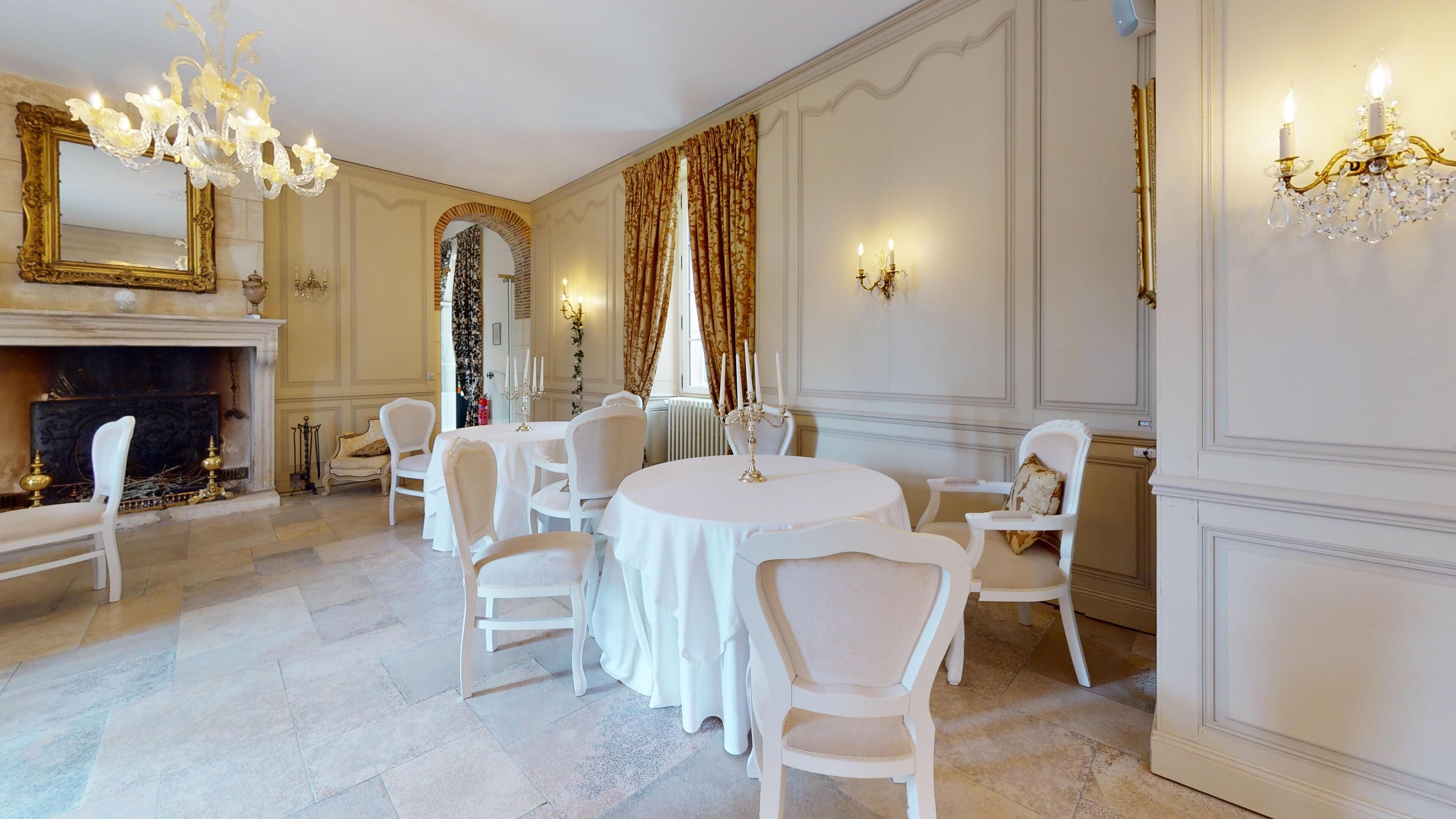 Een formele eetzaal met een open haard, witte stoelen, een ronde tafel en decoratieve wandklemmen.