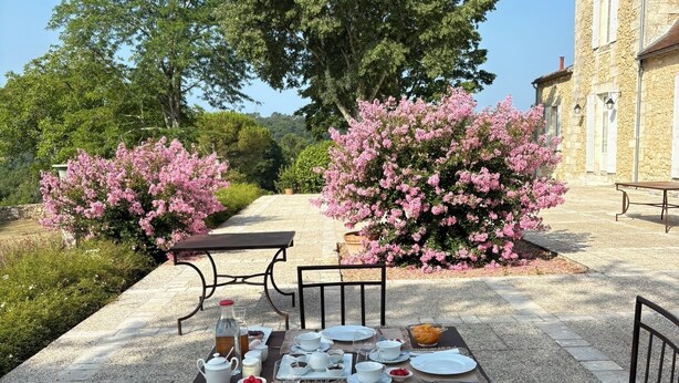 An outdoor dining area with a stone table set for a meal, surrounded by chairs and pink flowering bushes. - Château Ecrin de lumiere (Campsegret)