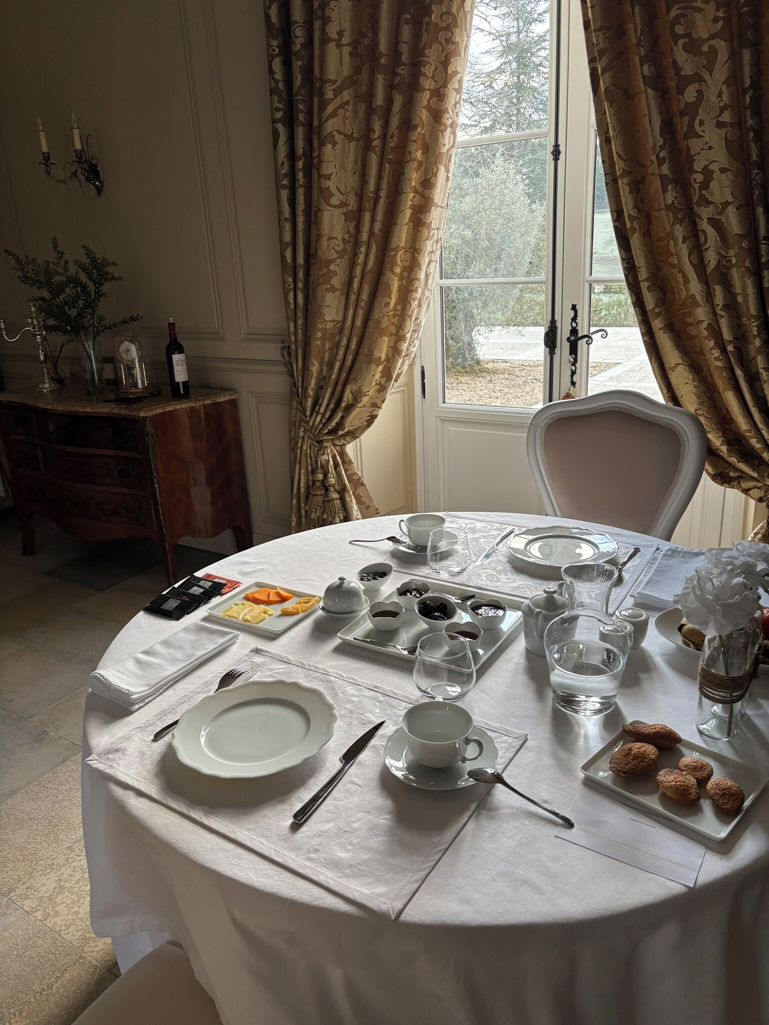 A dining table set with plates, cutlery, glasses, and a variety of food items, with a window and curtains in the background.