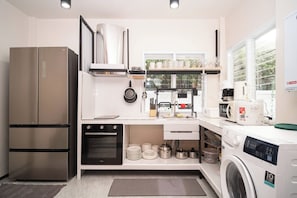 A modern kitchen with a stainless steel refrigerator, oven, and washing machine, white cabinetry, and a window providing natural light.