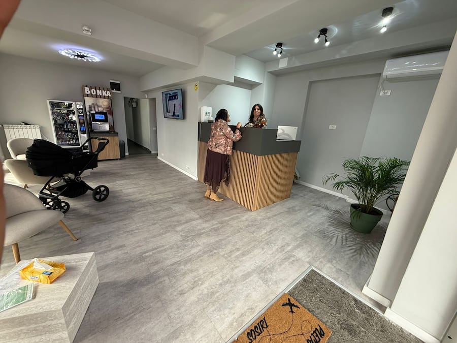 A reception area with a wooden reception desk, a refrigerator with beverages, a chair, a stroller, and a potted plant.
