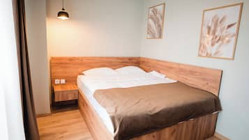 Un dormitorio con cabecera de madera, cama individual con ropa blanca, manta marrón, mesita de noche con lámpara y dos cuadros enmarcados.