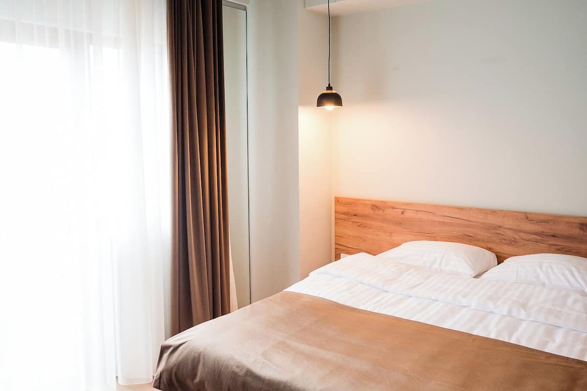 A modern bedroom with a wooden headboard, a large bed with white linens, a hanging pendant light, and sheer curtains.