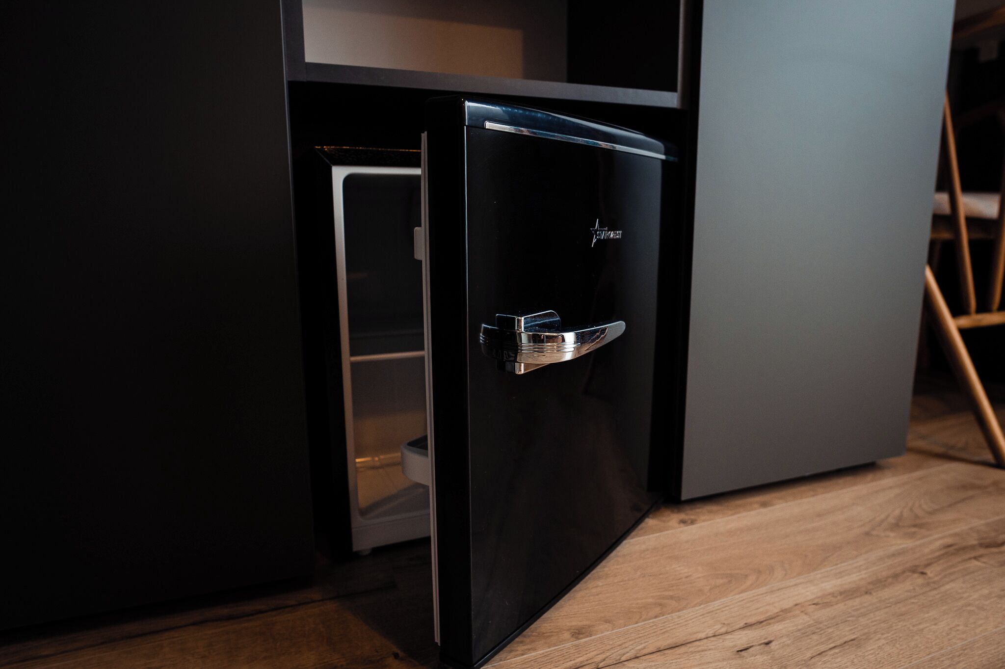 A compact, modern refrigerator with a metallic handle, situated in a kitchen with wooden flooring and dark cabinetry.