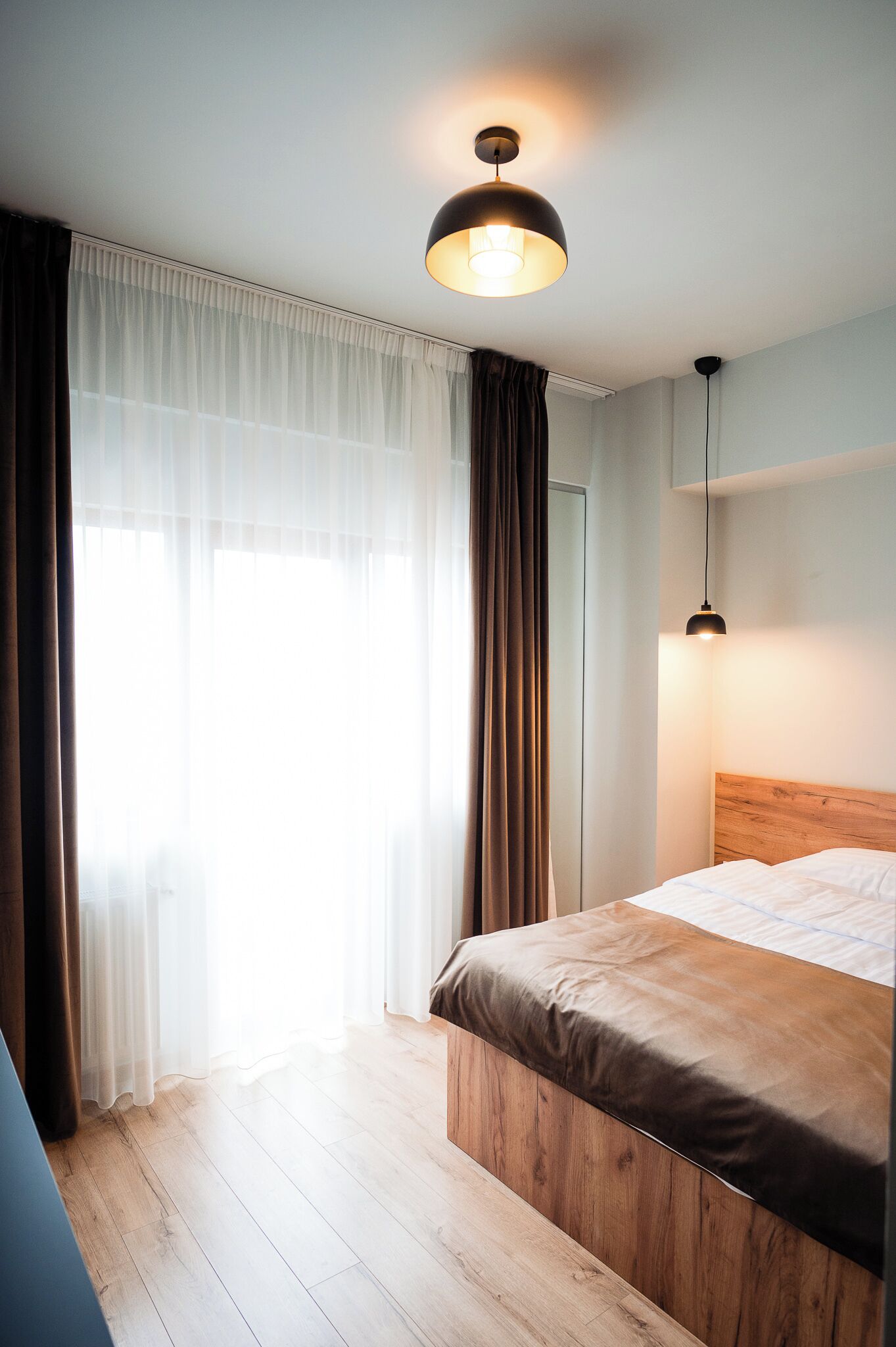 Camera da letto moderna con una testiera in legno, un letto grande, una lampada a sospensione e tende leggere che lasciano filtrare la luce naturale.