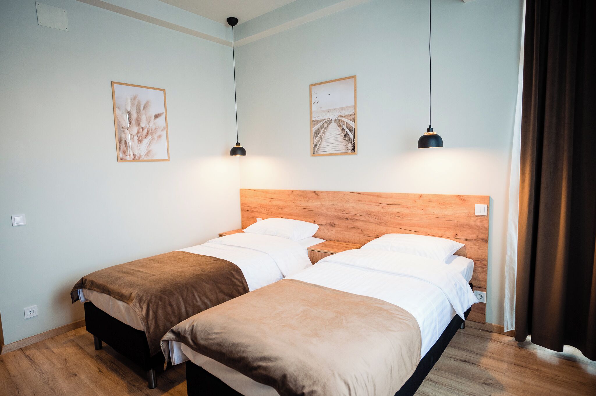Quarto com duas camas, cabeceira de madeira, duas luminárias pendentes e dois quadros na parede.