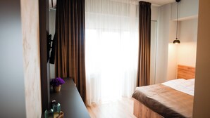A hotel room with a bed, a desk with a chair, a mirror, a window with curtains, and a hanging light fixture. - FLY & STAY AT GEORGE'S (Otopeni)