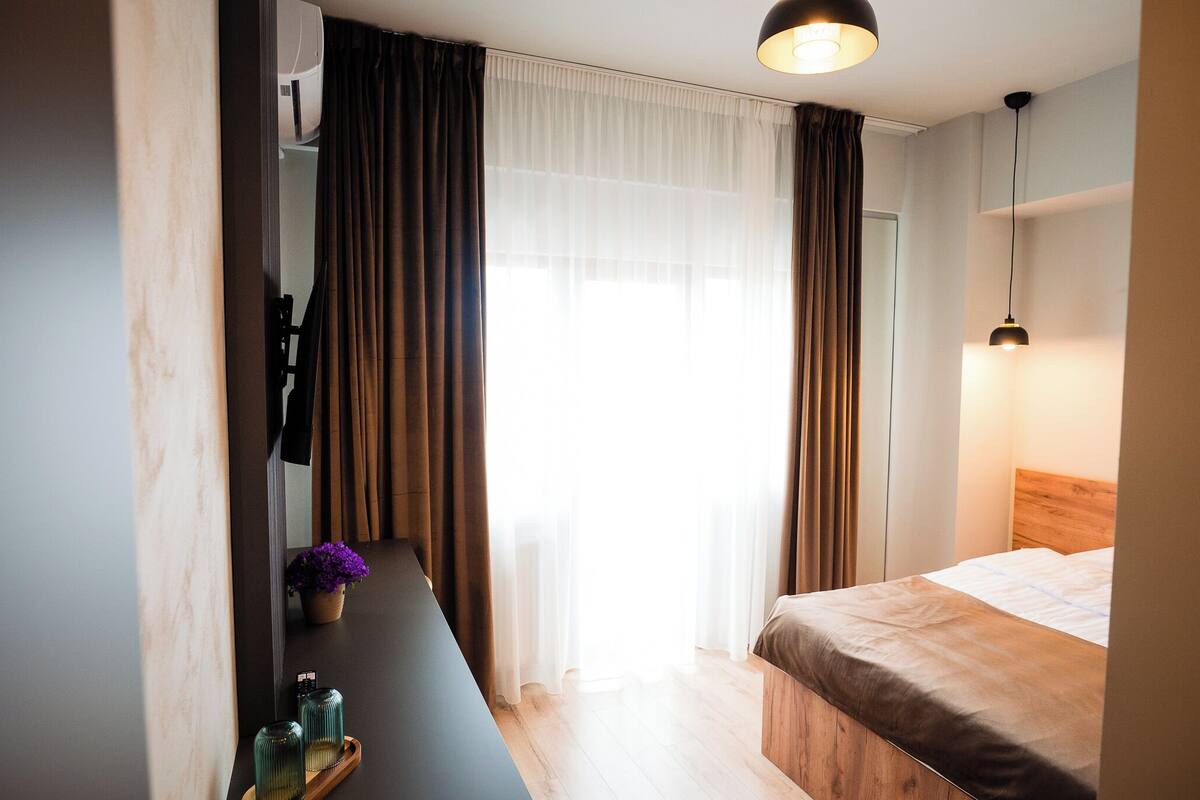 A hotel room with a bed, a desk with a chair, a mirror, a window with curtains, and a hanging light fixture.