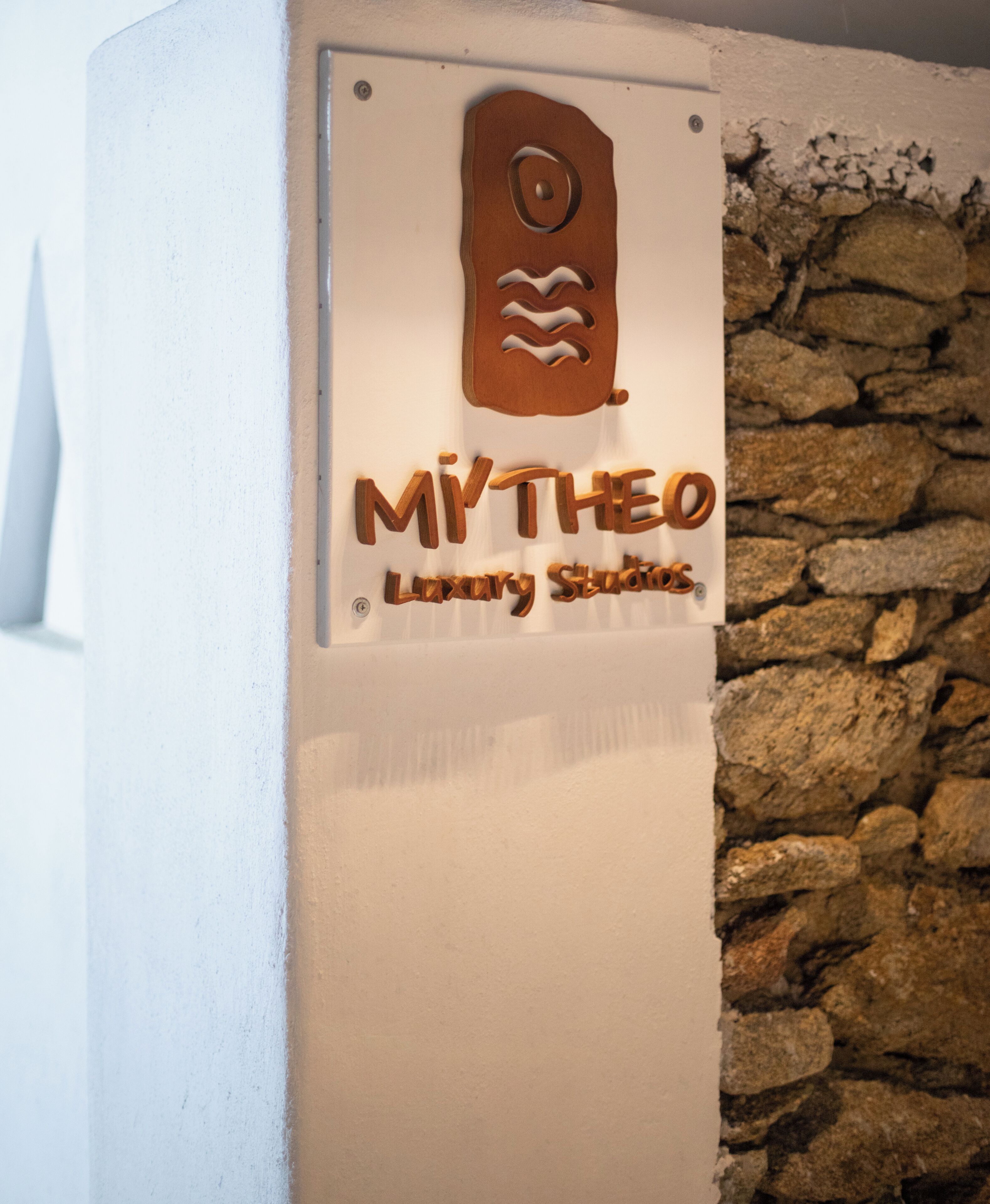 A sign with the name "MYTHEO Luxury Studios" mounted on a white wall with a stone wall background.