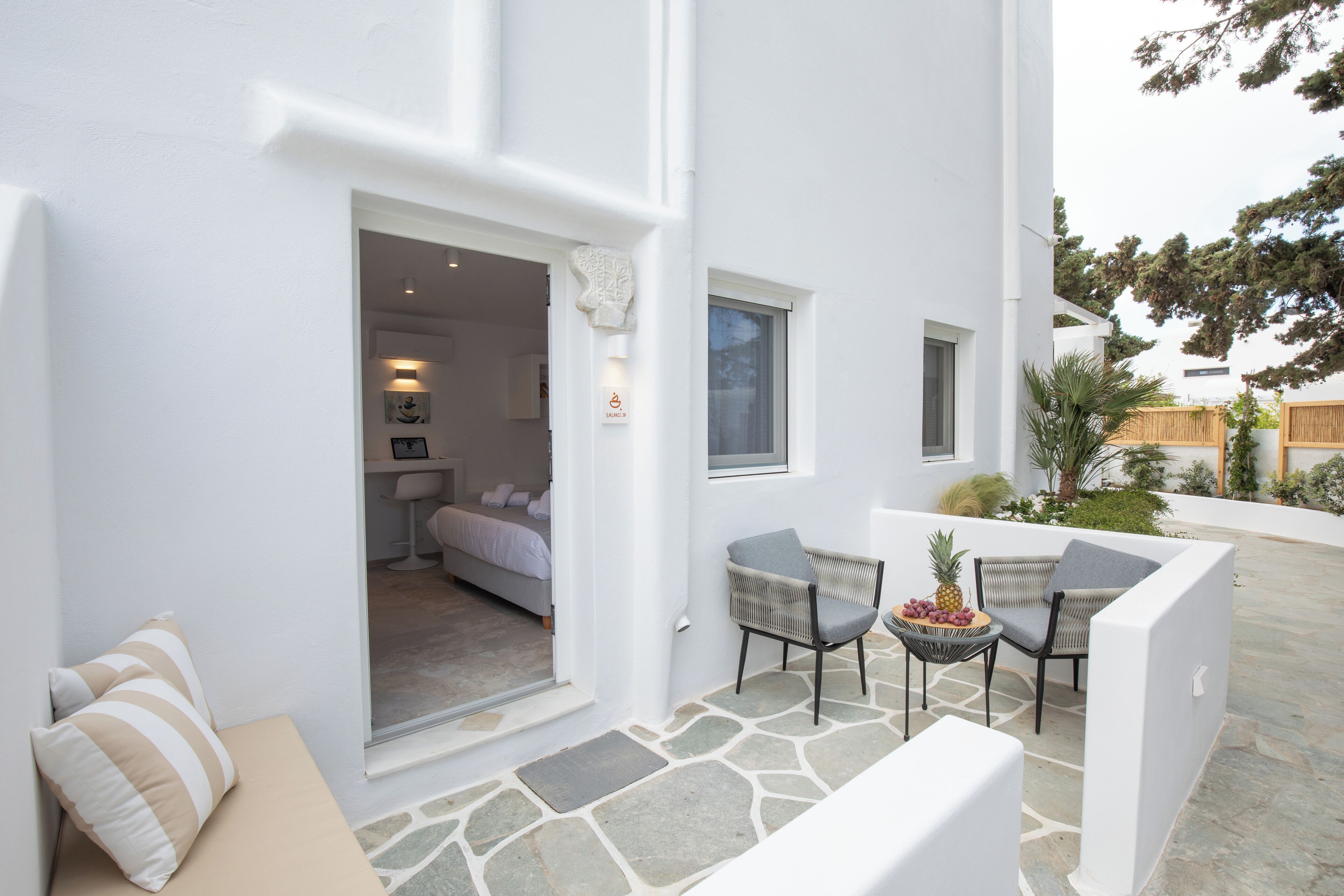 A white building with a stone patio, a bed, and a small dining area with chairs and a table.