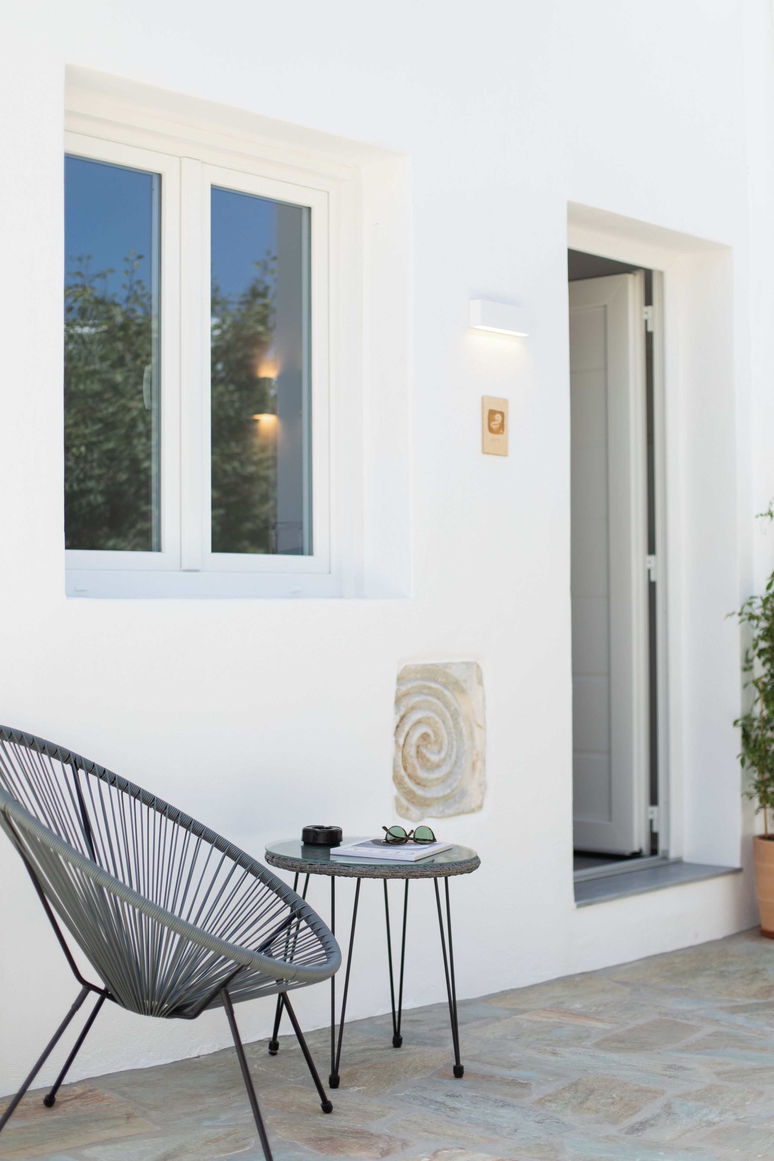 A white exterior wall with a window, a door, a small round table with a chair, and a potted plant.