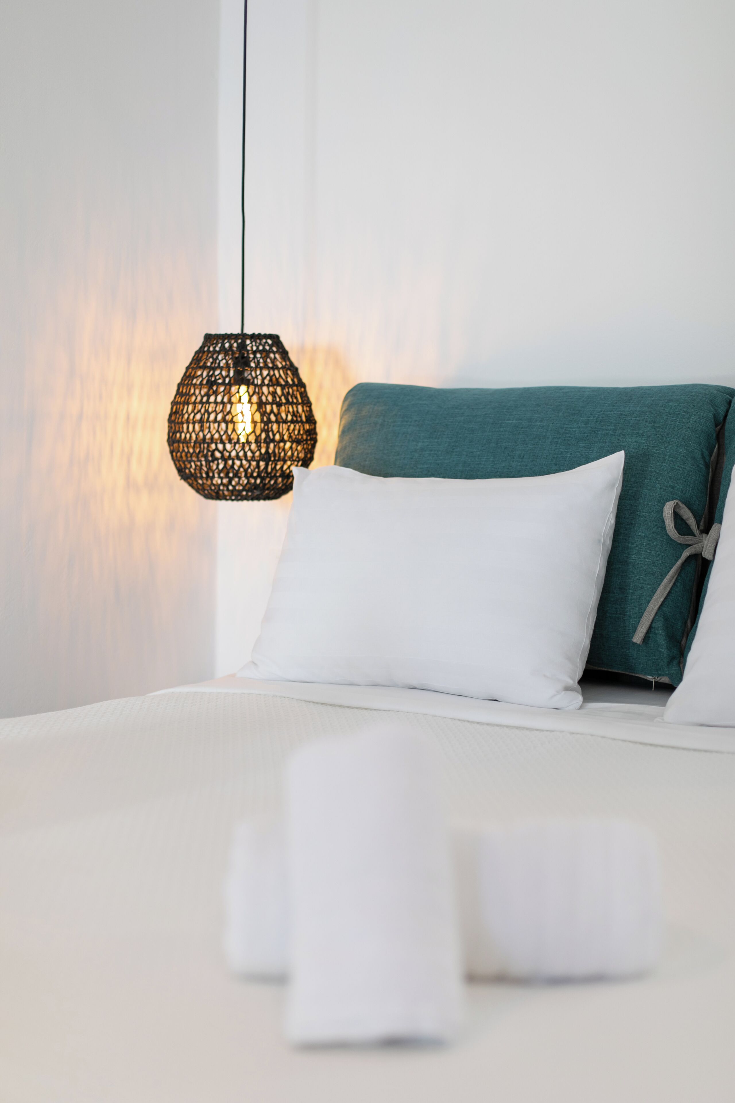 A neatly made bed with white linens and a teal pillow, a hanging woven light fixture, and a plain wall in the background.