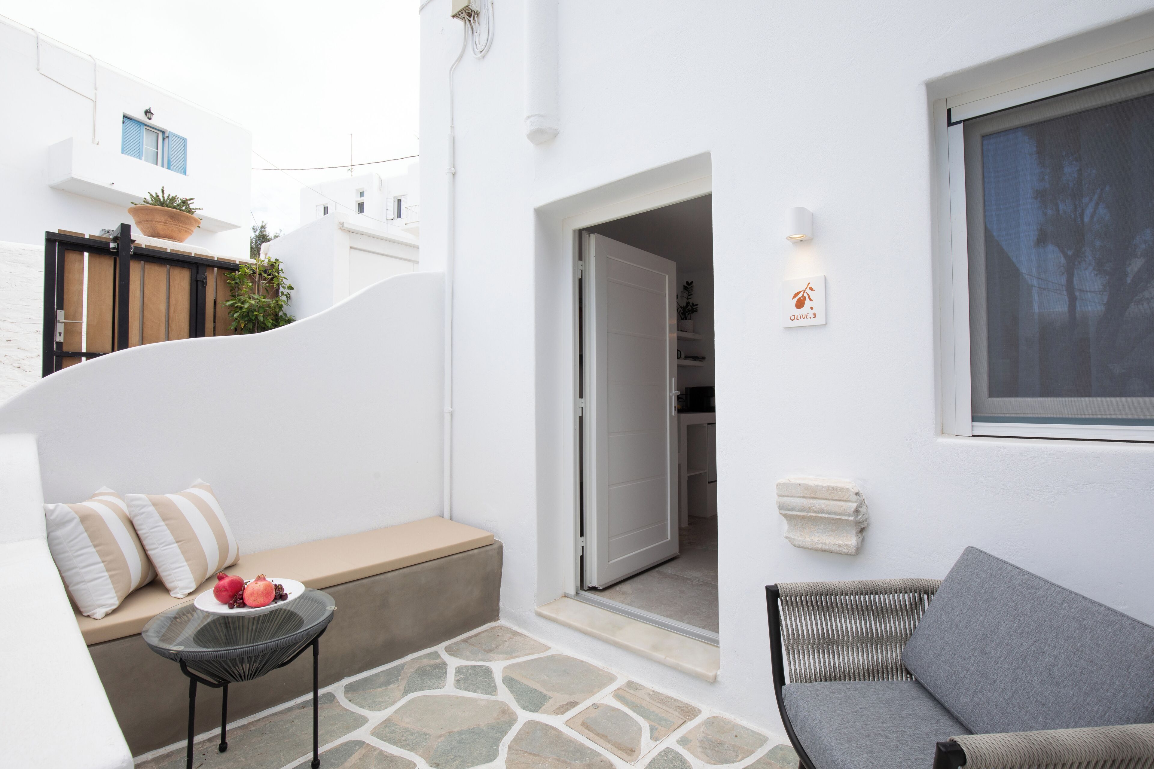 A white-walled room with a bench, a small table with fruit, a wicker chair, and a door leading to another room.