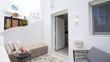 A white-walled room with a bench, a small table with fruit, a wicker chair, and a door leading to another room.