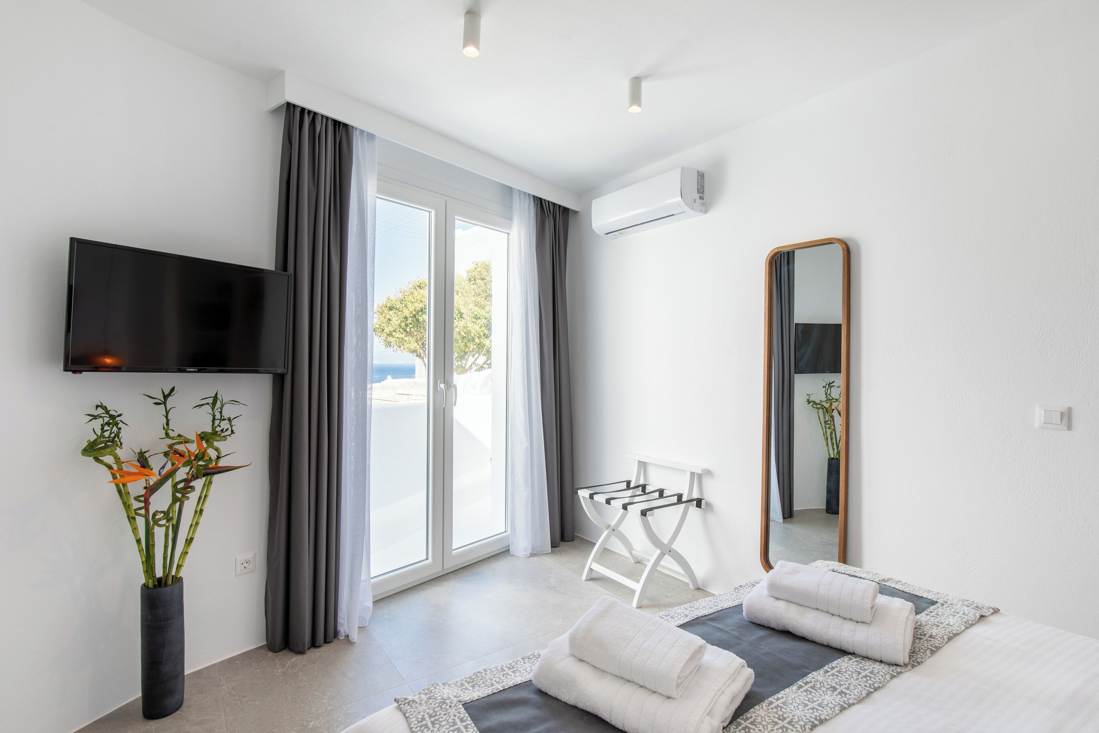 A modern hotel room with a large window, a flat-screen TV, a mirror, and a towel rack.