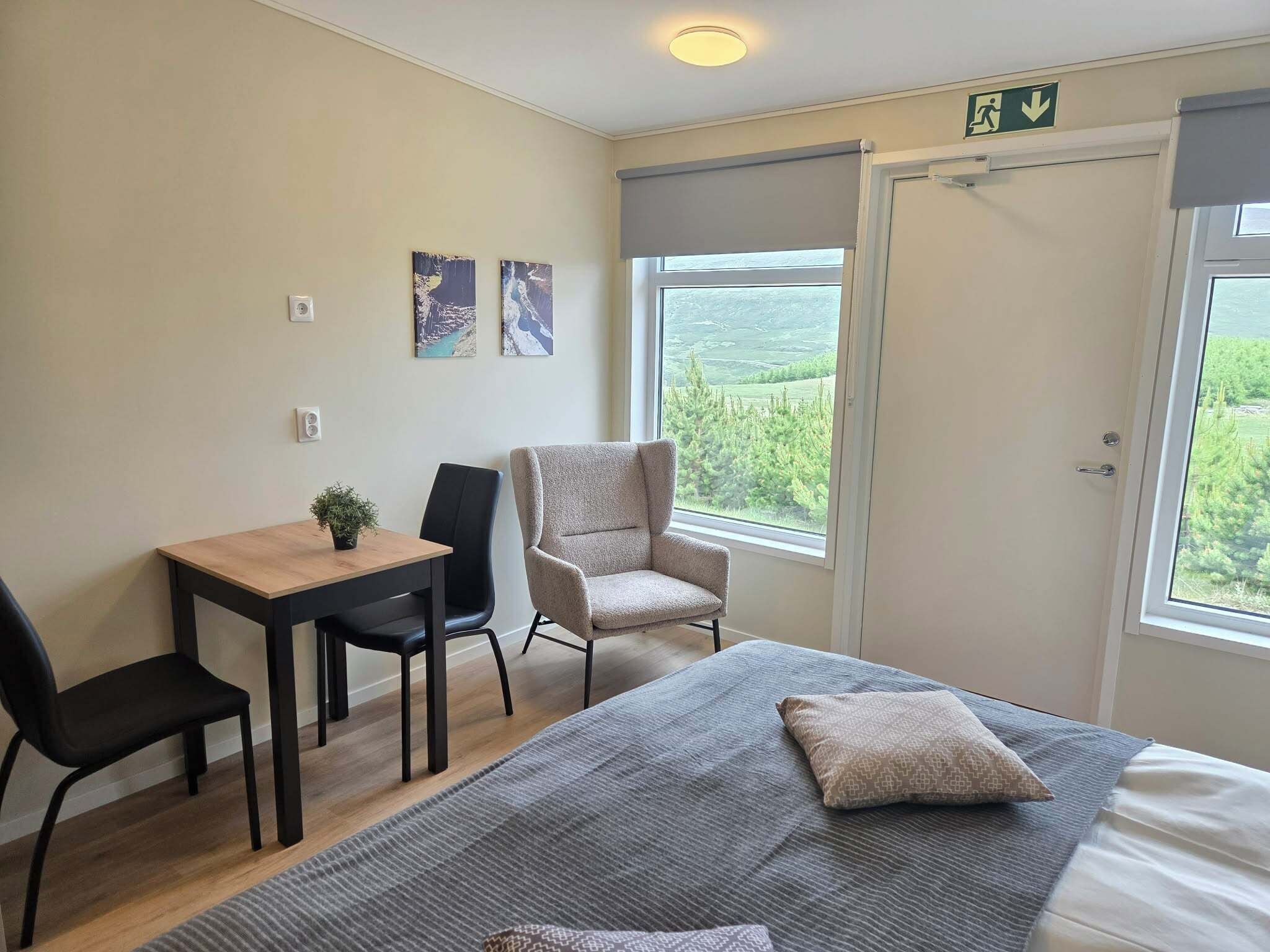 Un dormitorio con cama, escritorio, dos sillas, ventana con vistas y puerta con señal de salida de emergencia.