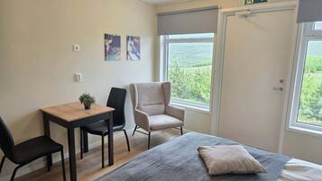 Un dormitorio con cama, escritorio, dos sillas, ventana con vistas y puerta con señal de salida de emergencia.