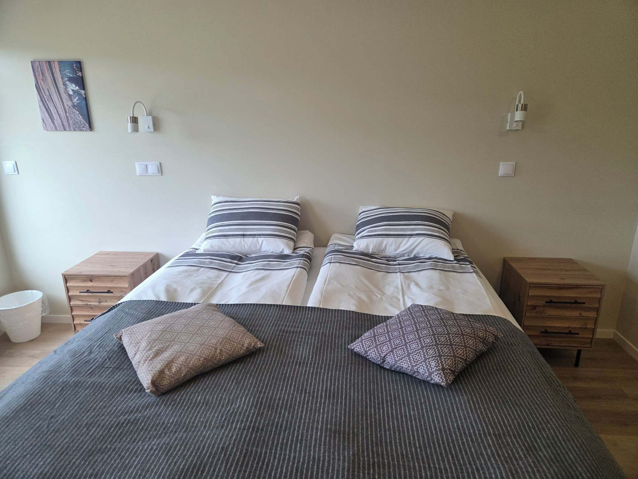 Cama doble con ropa de cama a rayas, dos mesitas de noche, una mesita de noche de madera y una lámpara fijada a la pared.
