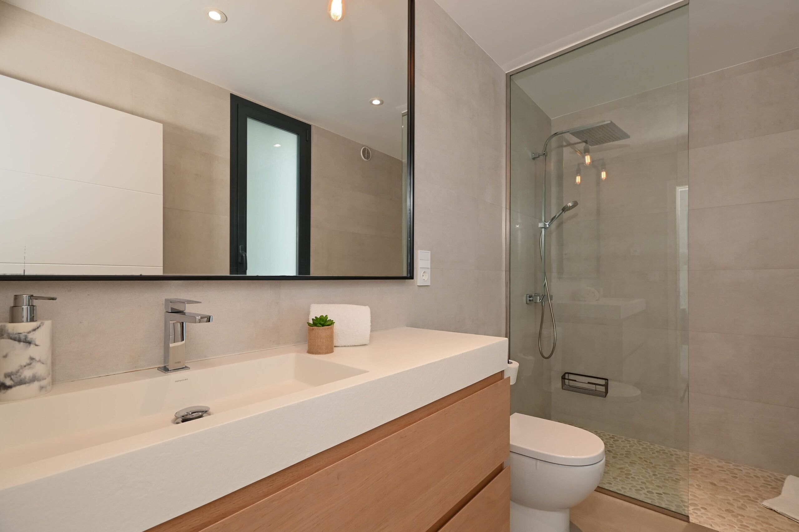 Une salle de bain moderne avec un grand miroir, un lavabo doté d’un distributeur de savon en marbre, des toilettes et une douche séparée par une paroi en verre.
