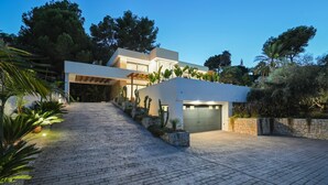 Une maison moderne avec une allée pavée menant à un garage, entourée d’une végétation luxuriante et d’arbres.