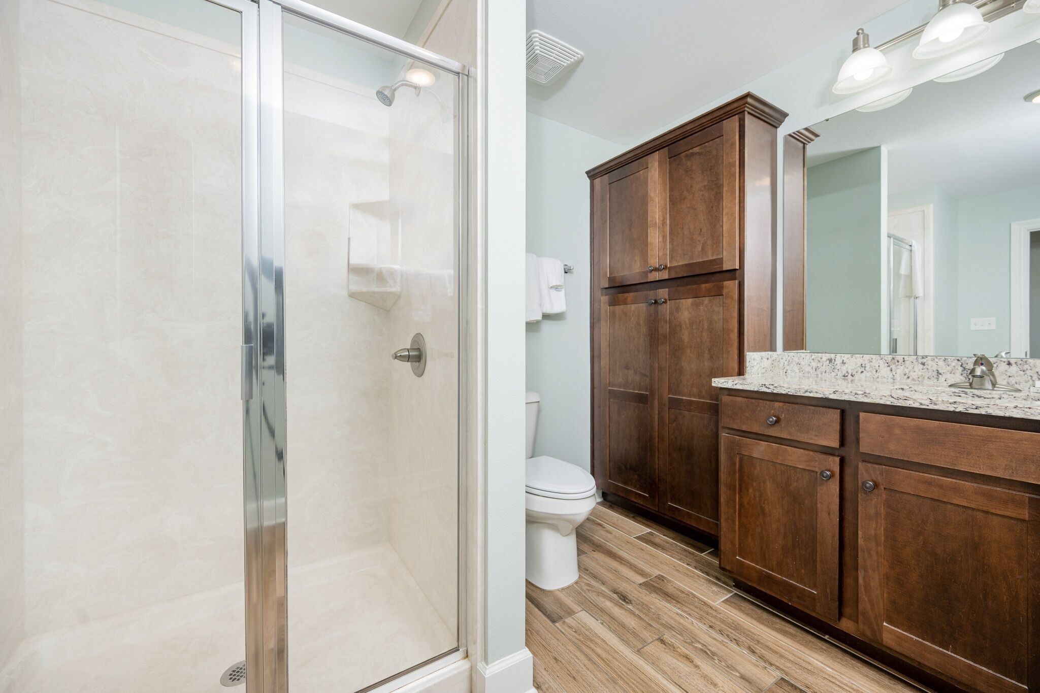 A bathroom with a glass shower enclosure, a toilet, wooden cabinets, and a marble countertop.