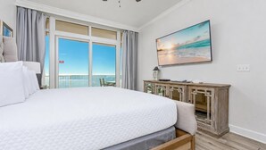 Une chambre d’hôtel avec un grand lit, une table de chevet en bois, un tableau représentant une plage et une vue sur l’océan par la fenêtre.