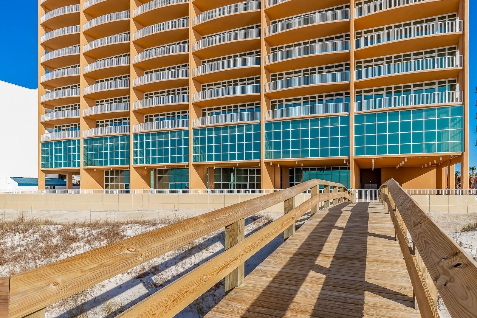 A multi-story hotel with balconies, a wooden boardwalk leading to the entrance, and a clear blue sky.