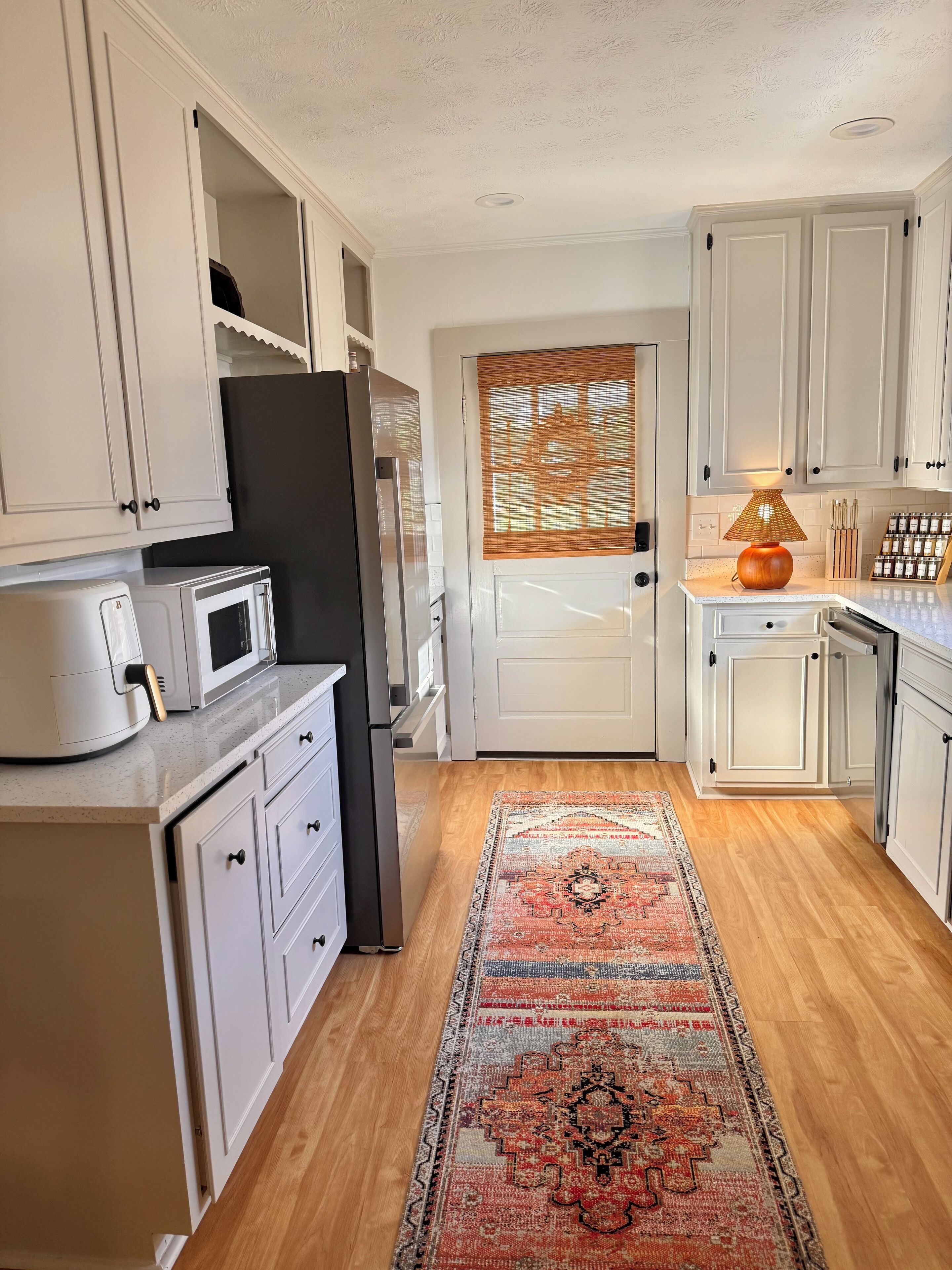 Une cuisine avec des meubles blancs, un four à micro-ondes, un réfrigérateur, un plancher en bois et un tapis à motifs.