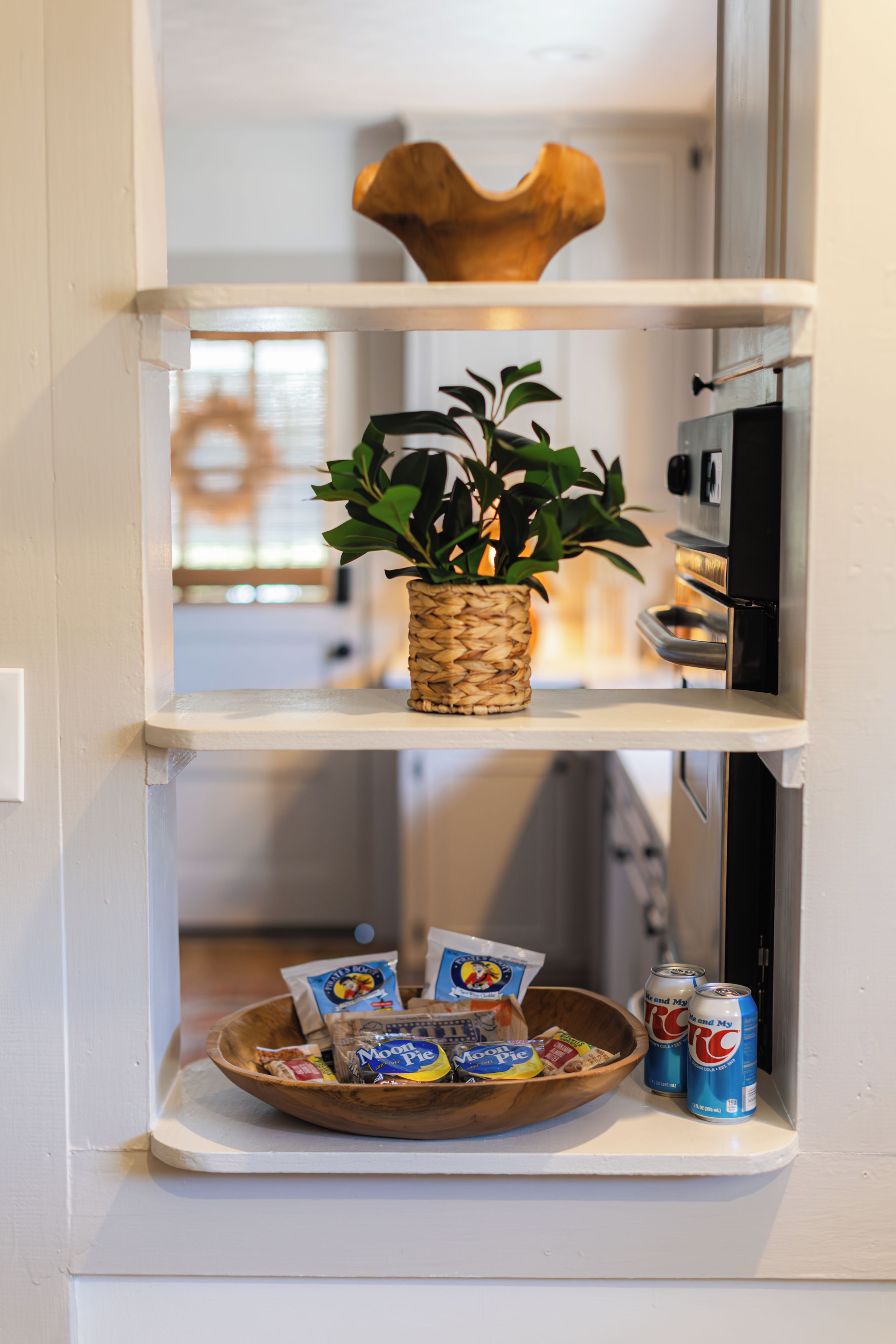 A kitchen shelf with a woven basket containing green plants, a wooden bowl with snacks and a can of soda, and a microwave above.