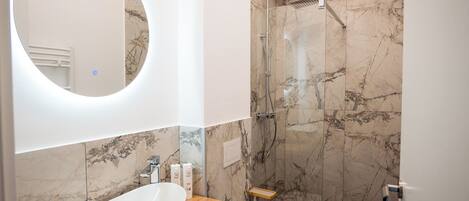 A modern bathroom with a large oval mirror, a white sink, a wooden countertop, a marble shower wall, and a small wooden shelf.