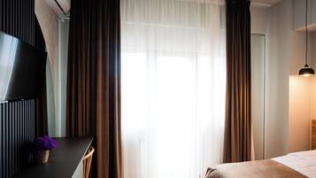Quarto de hotel com cama, escrivaninha com televisão, planta em vaso e janela com cortinas.
