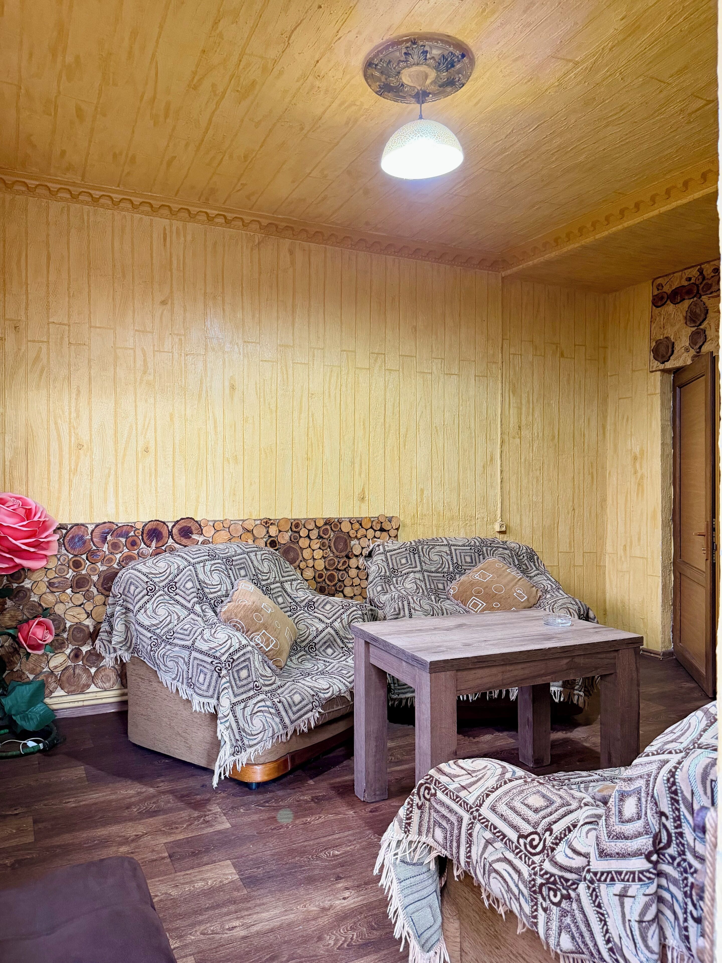 A cozy room with a wooden ceiling, a round ceiling light, a sofa with patterned cushions, a wooden coffee table, and a wall decorated with wooden logs.