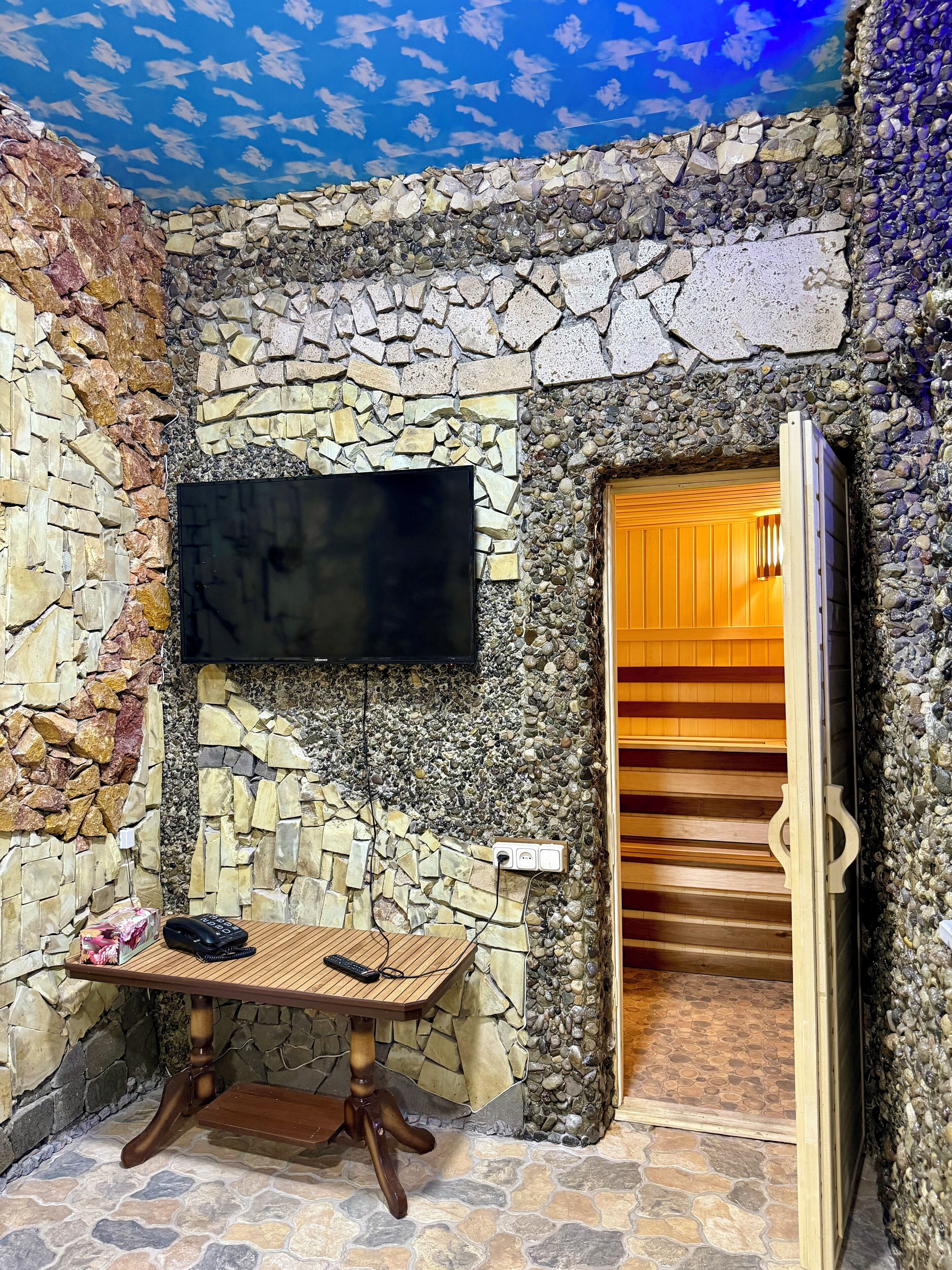 Ein Raum mit Steinwänden, an der Wand ist ein Flachbildfernseher angebracht, ein Holztisch mit einer Fernbedienung und eine Holztür, die in einen anderen Raum führt.