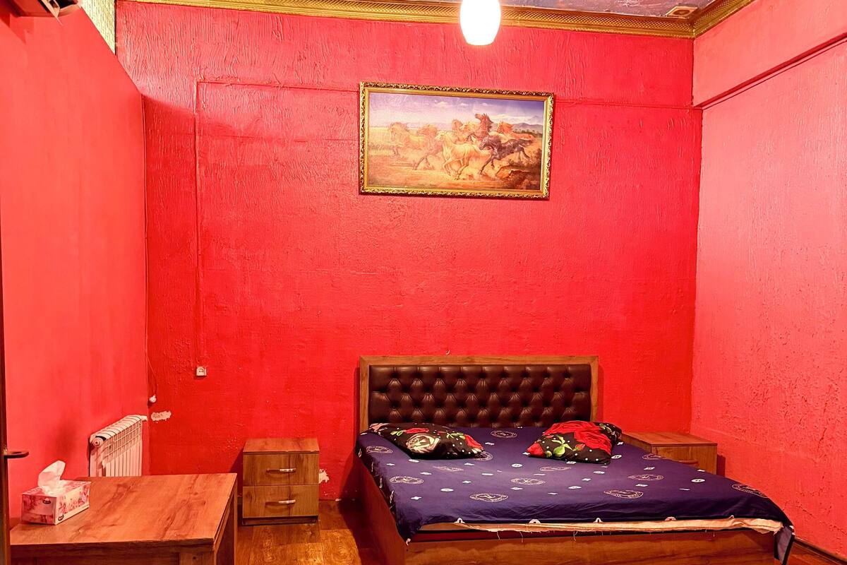 Quarto com parede vermelha, cama de madeira, escrivaninha de madeira e radiador.