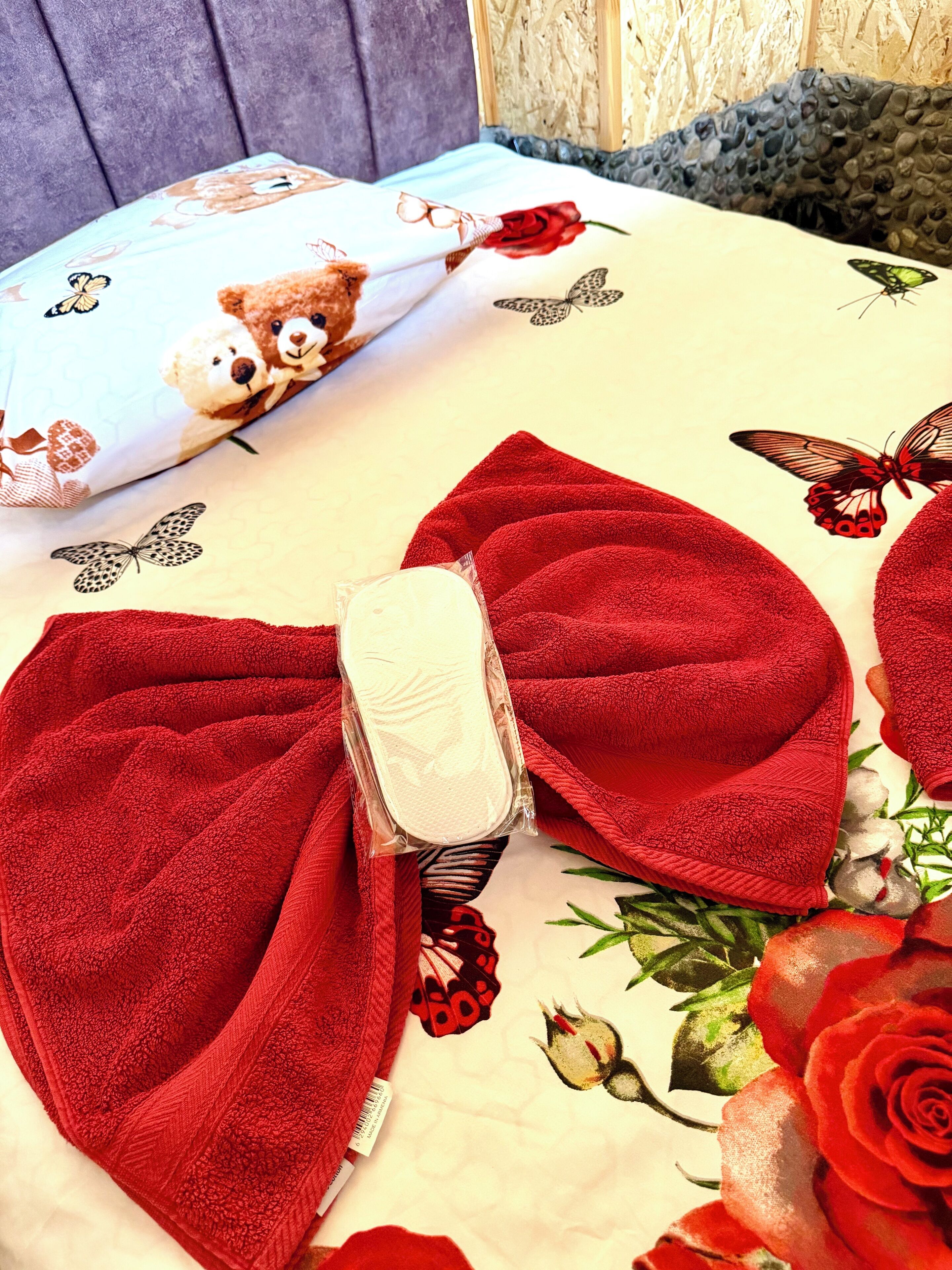 A bed with a floral bedspread, red towels, and a teddy bear pillow.