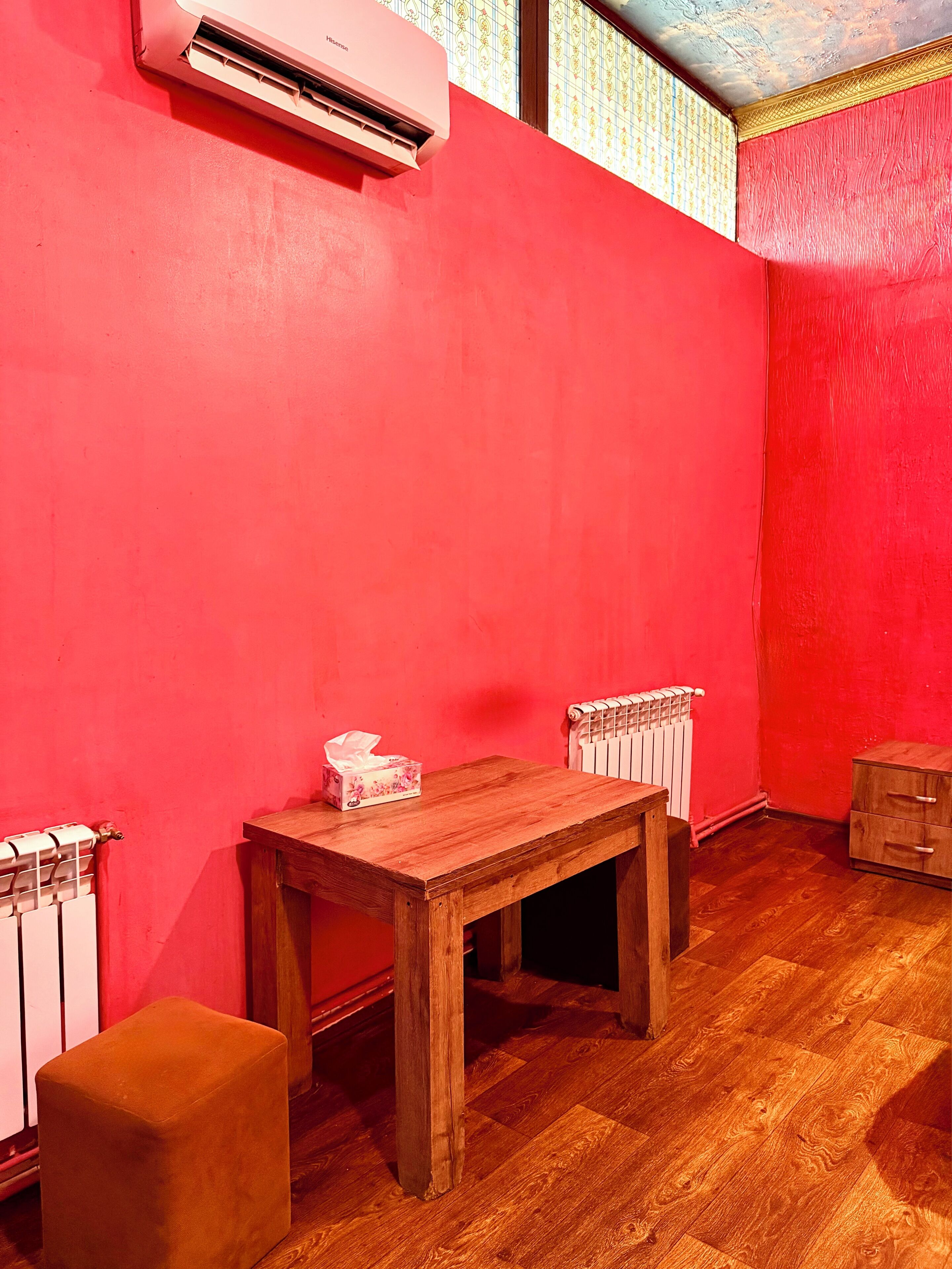 Een kamer met een rode muur, een houten tafel, een radiator en een klein houten kastje.