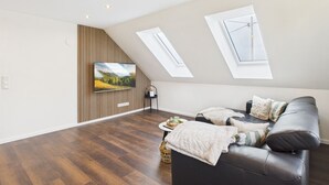 A modern living room with a flat-screen TV mounted on a wooden panel wall, a black leather sofa, a small side table with decorative items, and a skylight above the sofa. - Holiday apartment in the Eifel - for those seeking peace and quiet and nature lovers directly on the cycle path (Arzfeld)