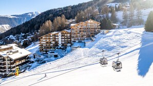 A ski resort with multiple buildings, including a large hotel with multiple floors and balconies, surrounded by snow-covered mountains and trees. - Victoria II E7 - 10 pers - at the foot of the slopes (Haute-Nendaz)