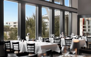 A restaurant with multiple tables, each set with white tablecloths and chairs, and large windows offering a view of palm trees and buildings.