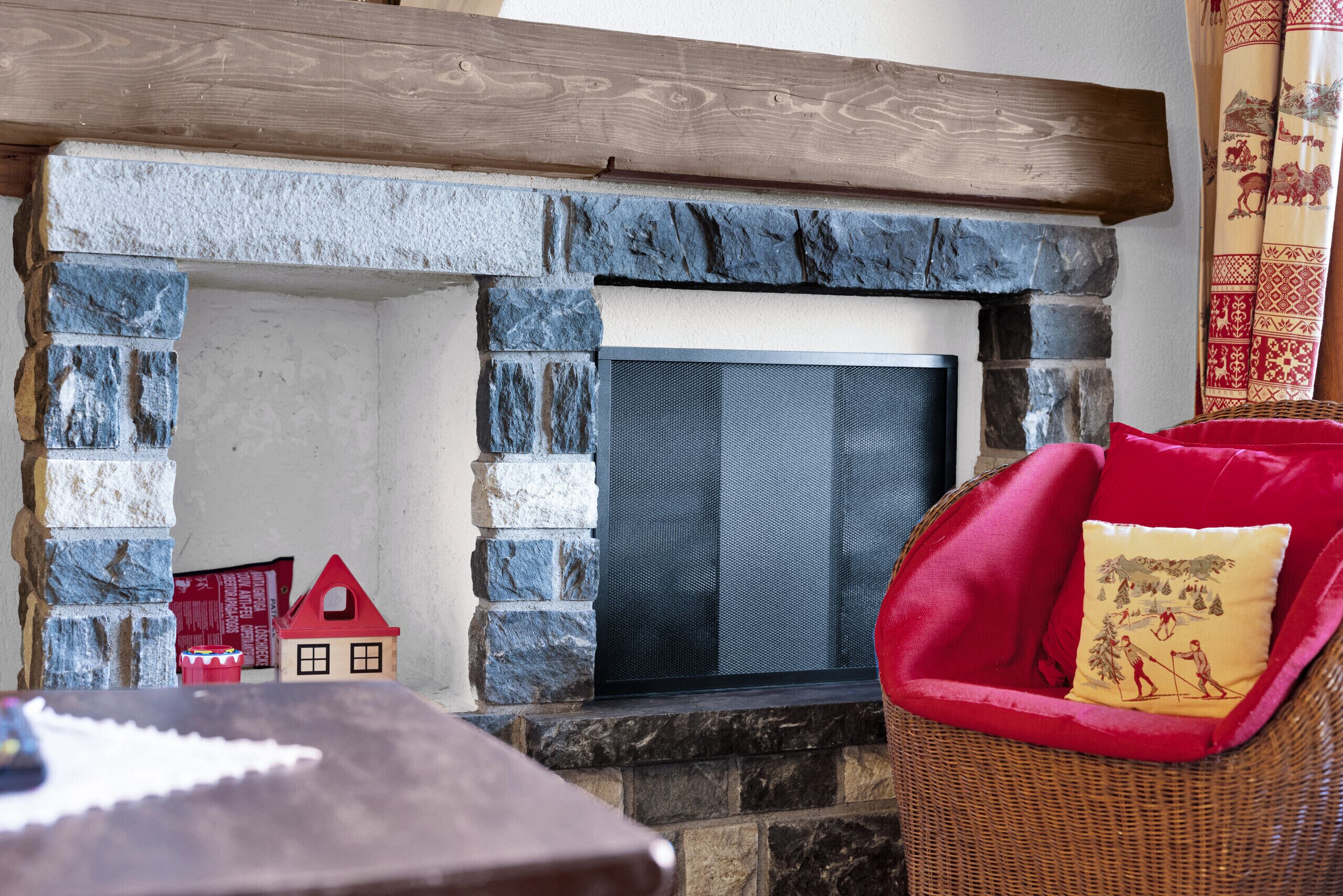A stone fireplace with a metal screen, a red chair with a cushion, and a small red house model.